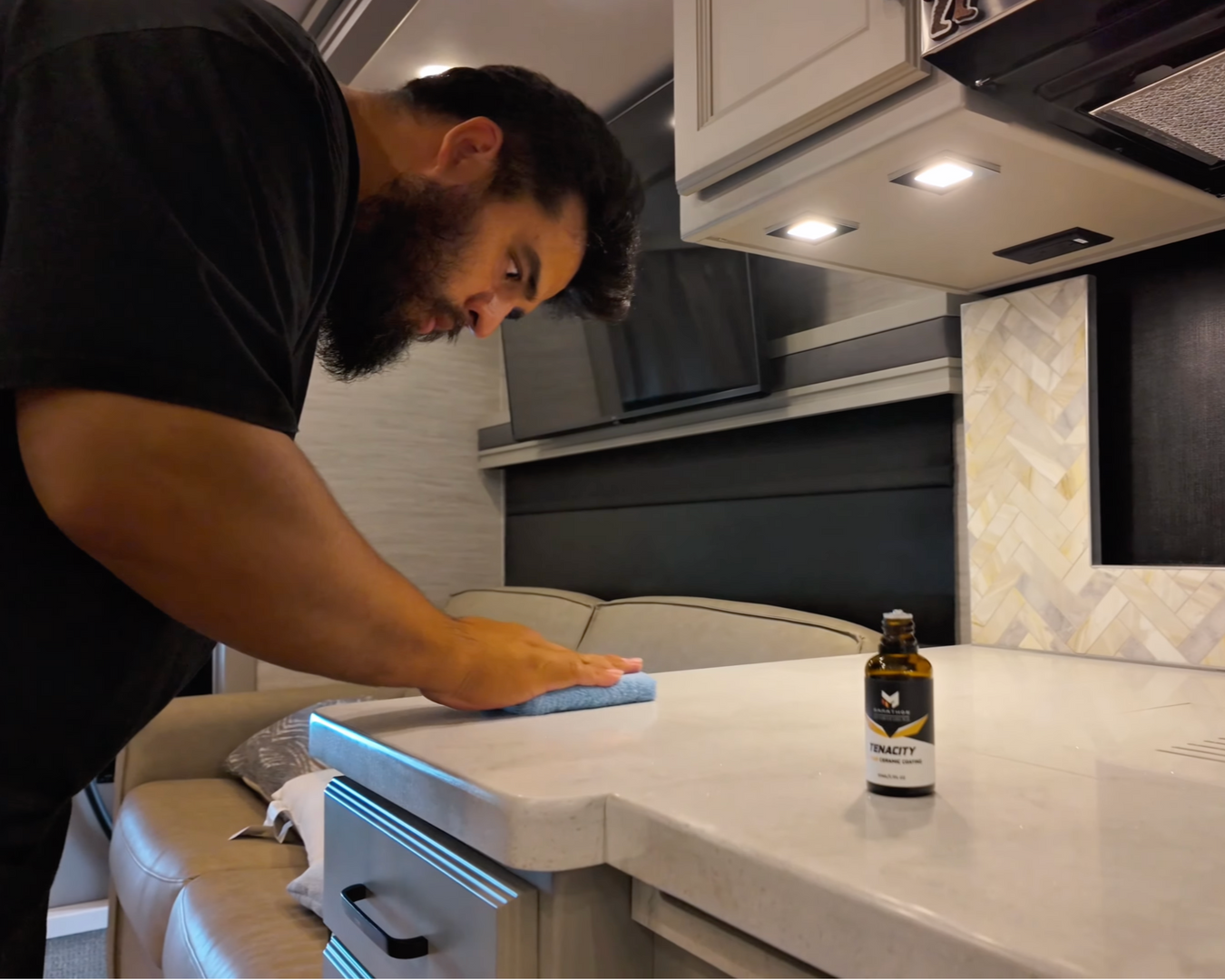 Person wiping a white kitchen countertop beside a small bottle under cabinet lights