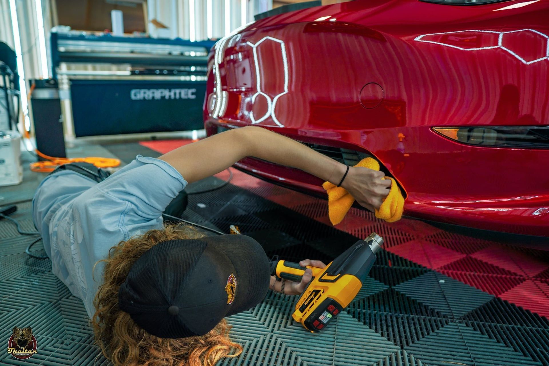 A woman is laying on the floor working on a red car.