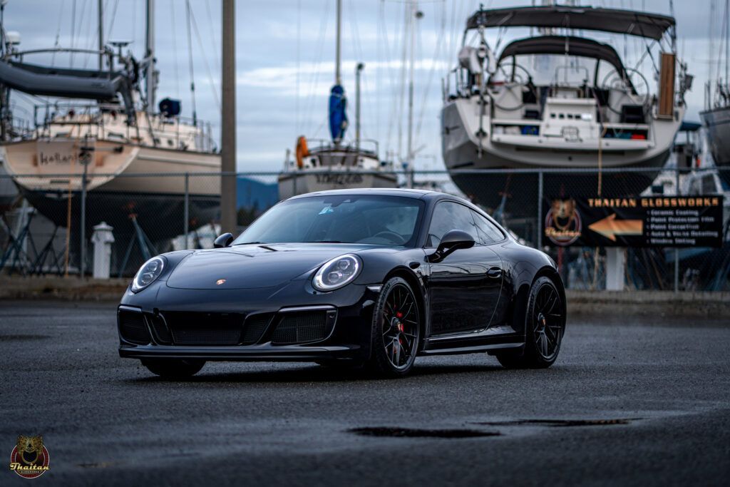 A black sports car is parked in front of boats in a marina.