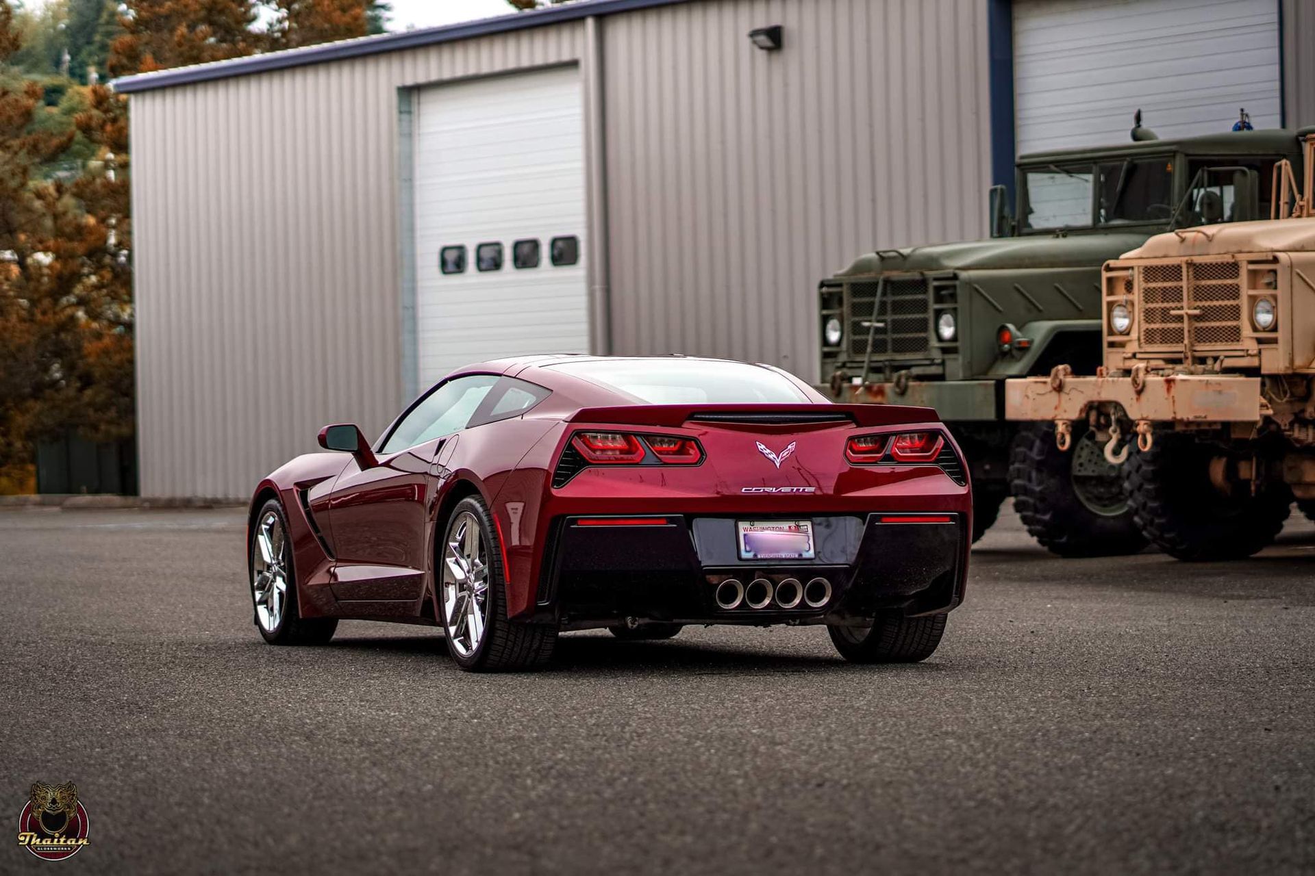 A red sports car is parked in front of a garage next to military vehicles.