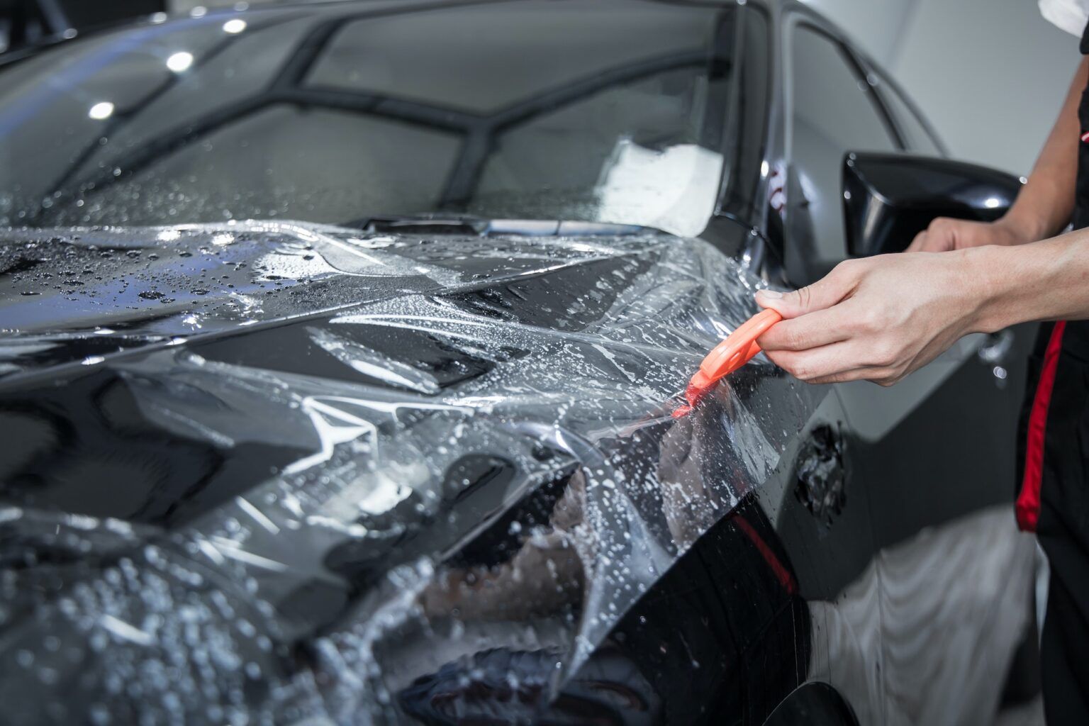 A person is wrapping a black car with a clear plastic film.