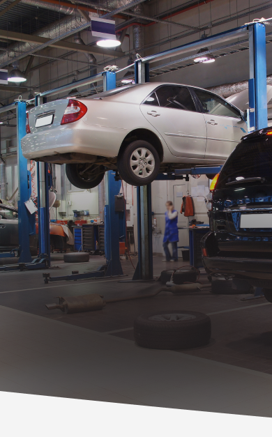 Silver car on a lift in a repair shop. A person stands nearby. Tools and another vehicle are visible. | Prestige Tire & Auto