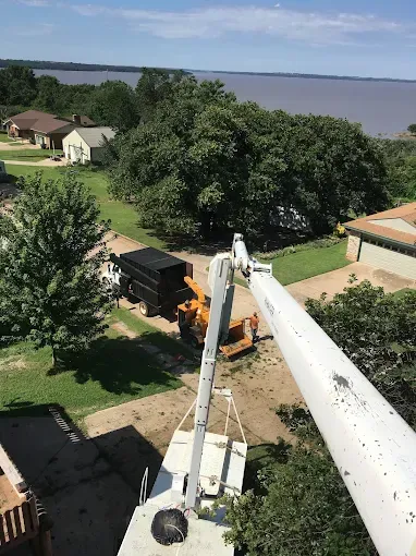 A high-angle view of a boom lift arm extending over a residential yard near Kaw Lake with a wood chipper and a trailer.