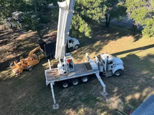 A high-angle view shows a white boom truck with its stabilizers extended and boom raised, parked near a wood chipper.