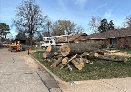 A pile of cut tree logs sits on a Ponca City front lawn near a tree removal truck and a wood chipper.