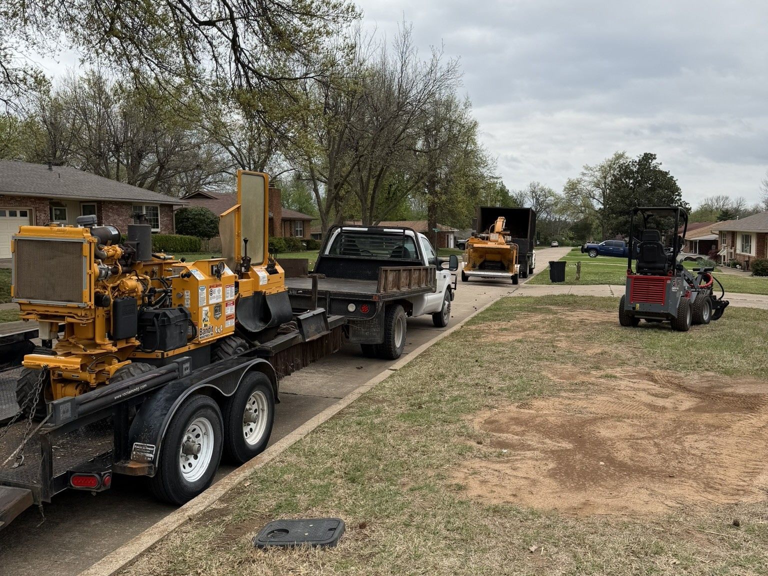 A yellow stump grinder sits on a trailer hitched to a truck, with another machine parked on the lawn of a residential area.