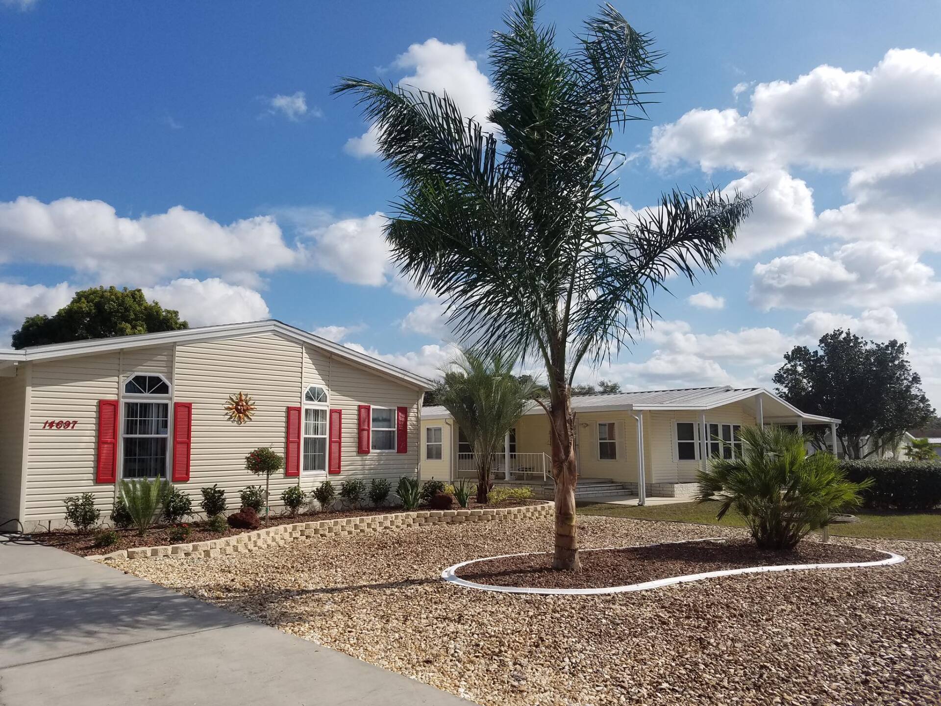 Houses with red shutters and a palm tree under a blue sky, surrounded by brown stones.