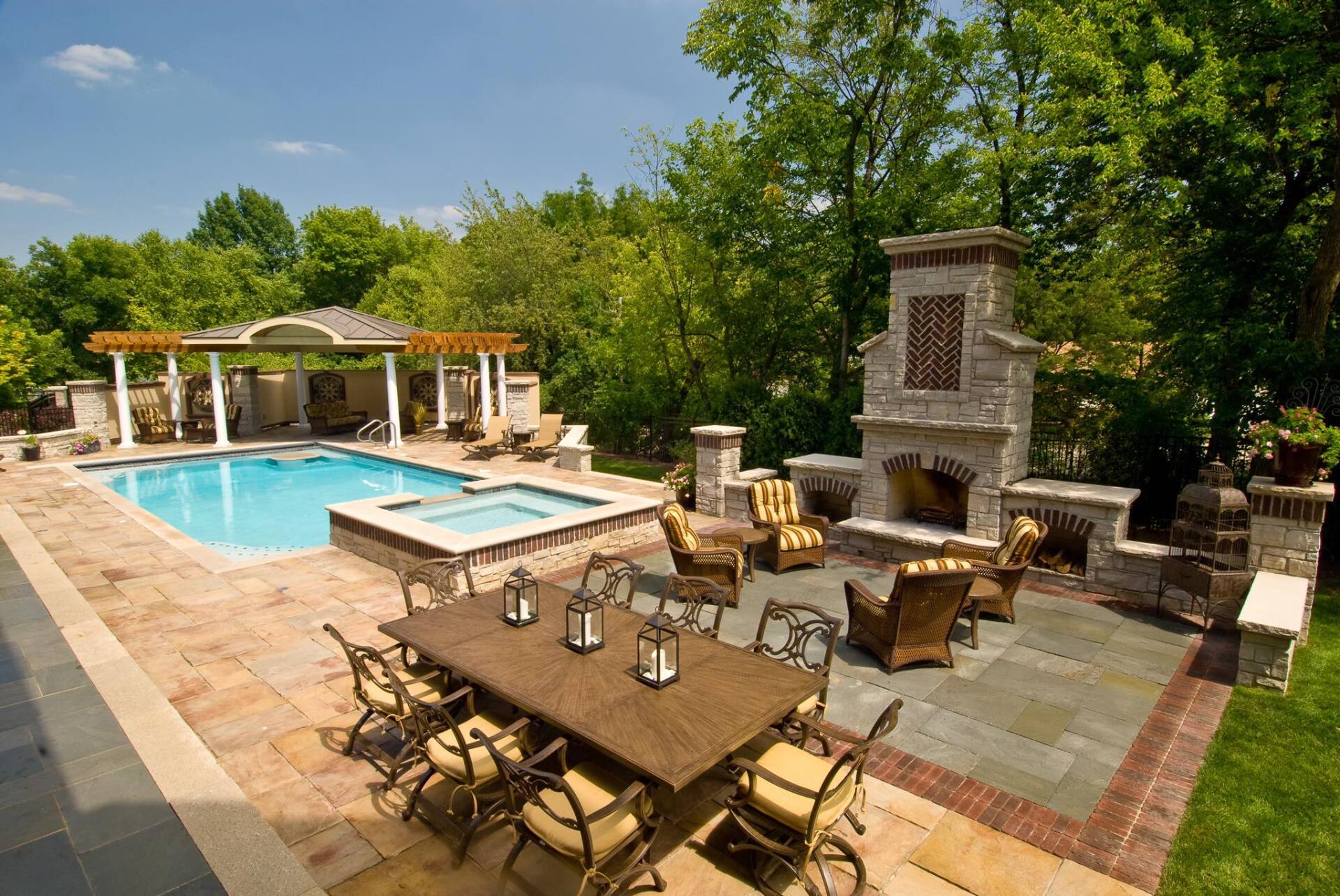 Outdoor patio with pool, hot tub, fireplace, dining area, and arbor; lush trees in the background.