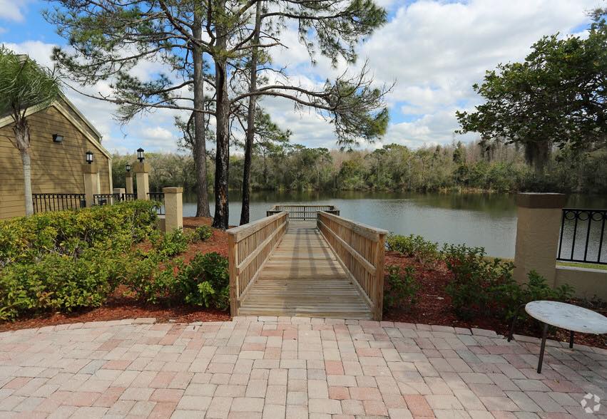Wooden pier extends over a lake with trees and a building in the background. Red brick patio and table in the foreground.