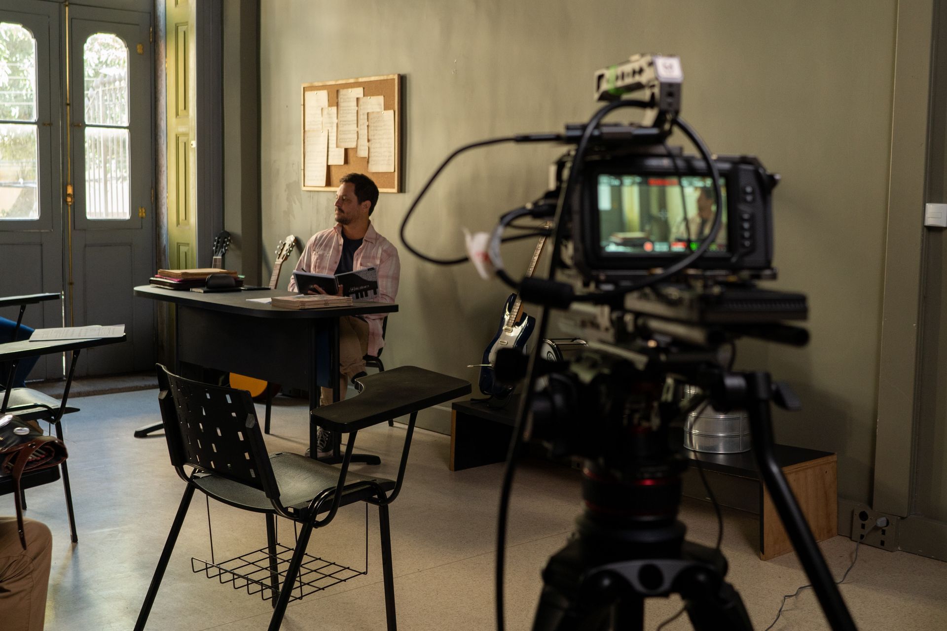 A man is sitting at a desk in front of a camera in a classroom.