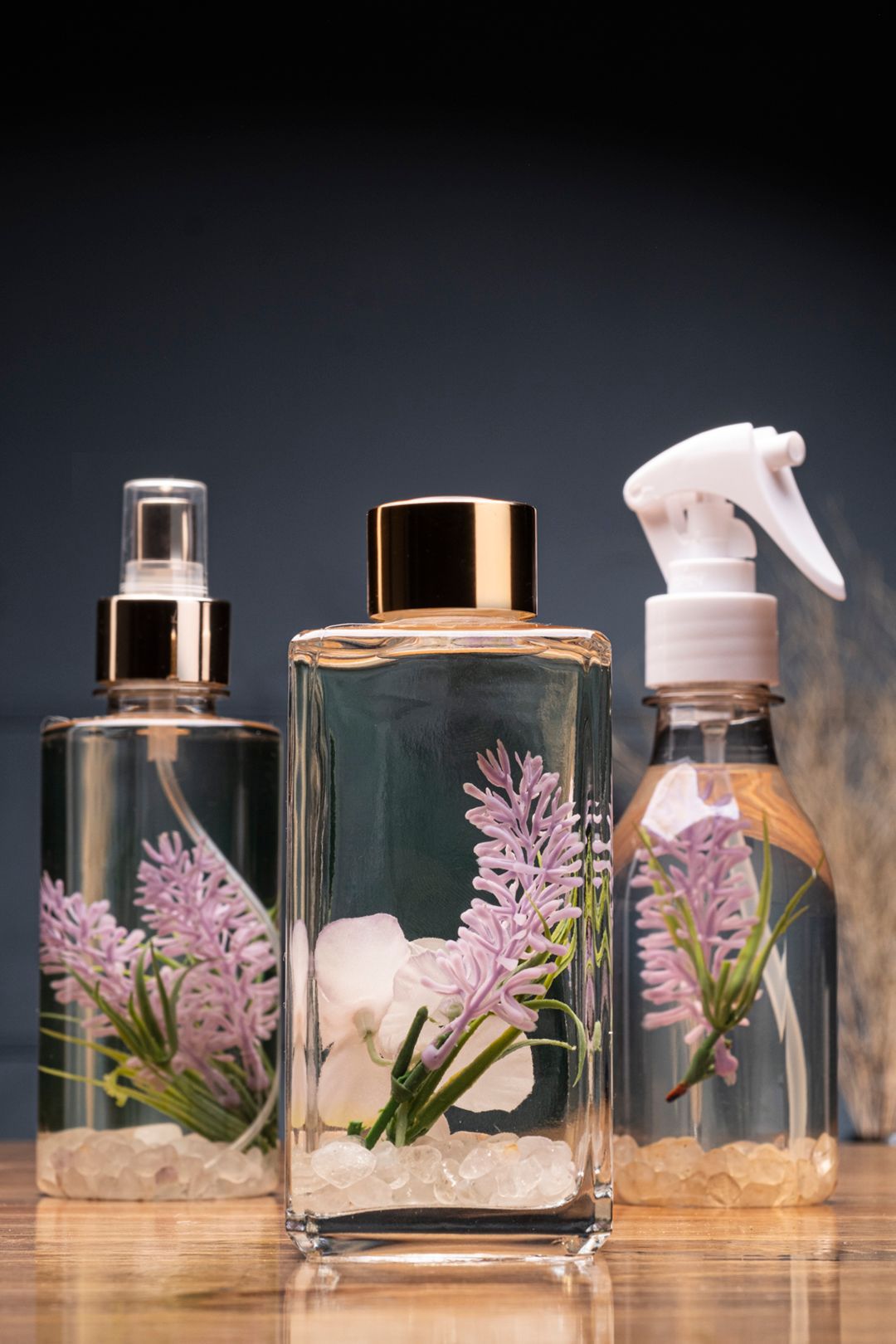 Três garrafas de vidro transparente com raminhos de lavanda e pedrinhas brancas, sobre uma superfície refletora.