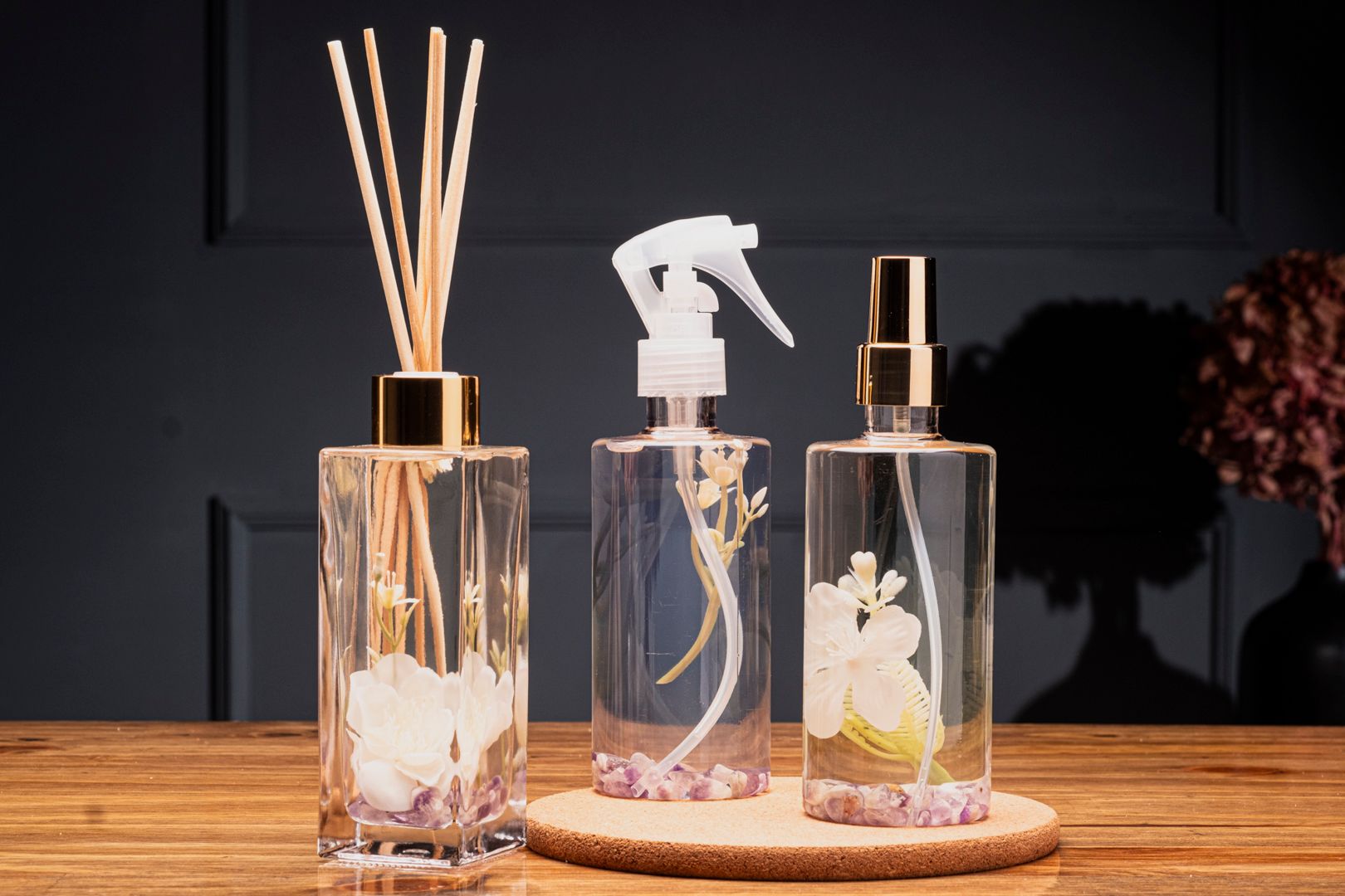 Três garrafas de vidro decorativas com detalhes dourados e flores secas sobre uma mesa de madeira.