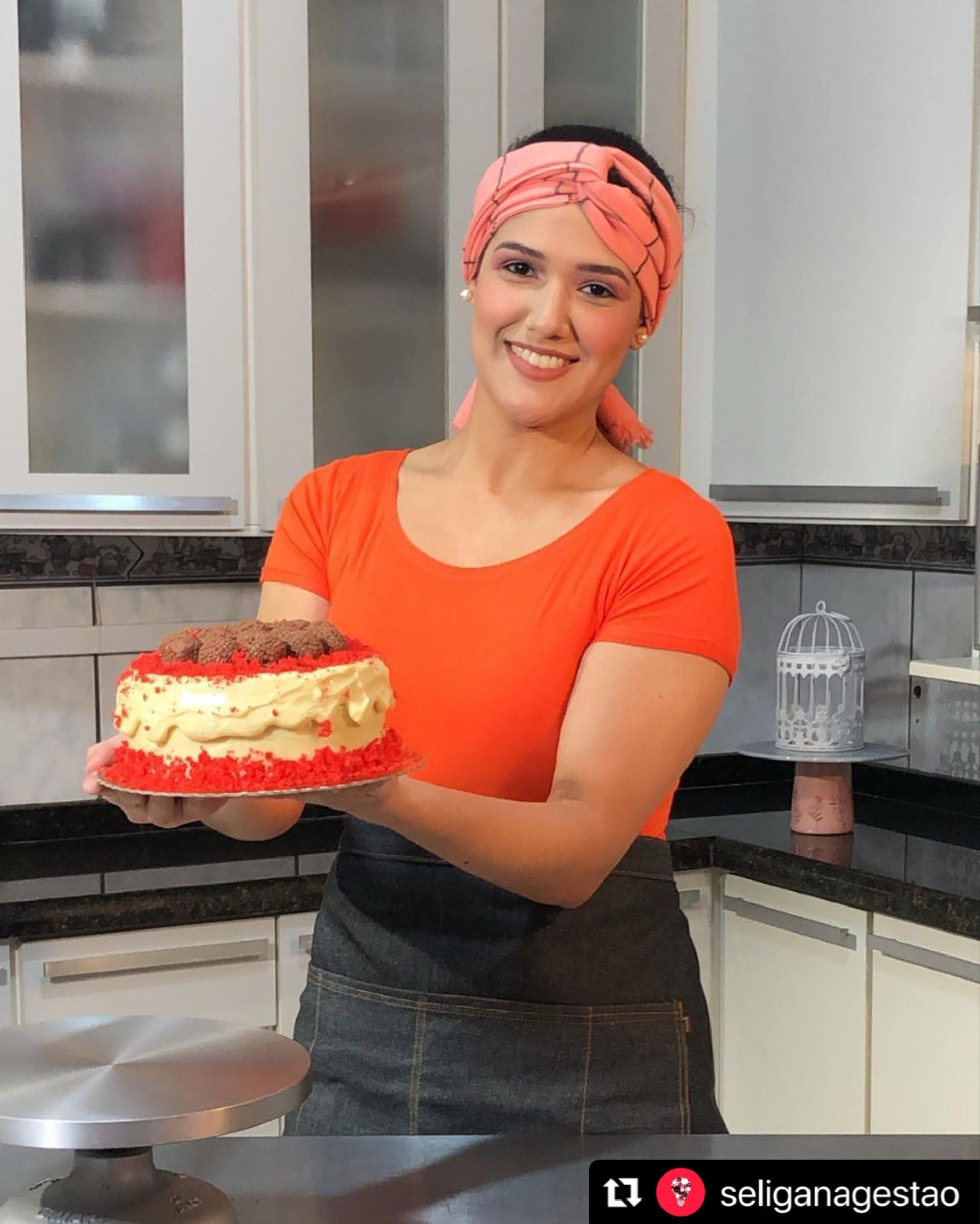 Mulher sorridente segurando um bolo de veludo vermelho em uma cozinha, vestindo blusa laranja e um turbante.