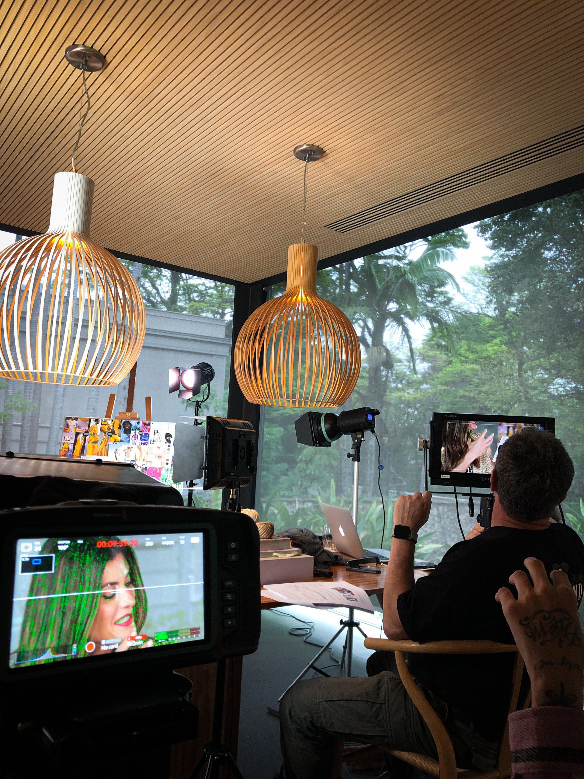 Cenário de filmagem: Mulher de cabelo verde em frente a um monitor, o diretor observa, luzes fortes no teto, janela grande.
