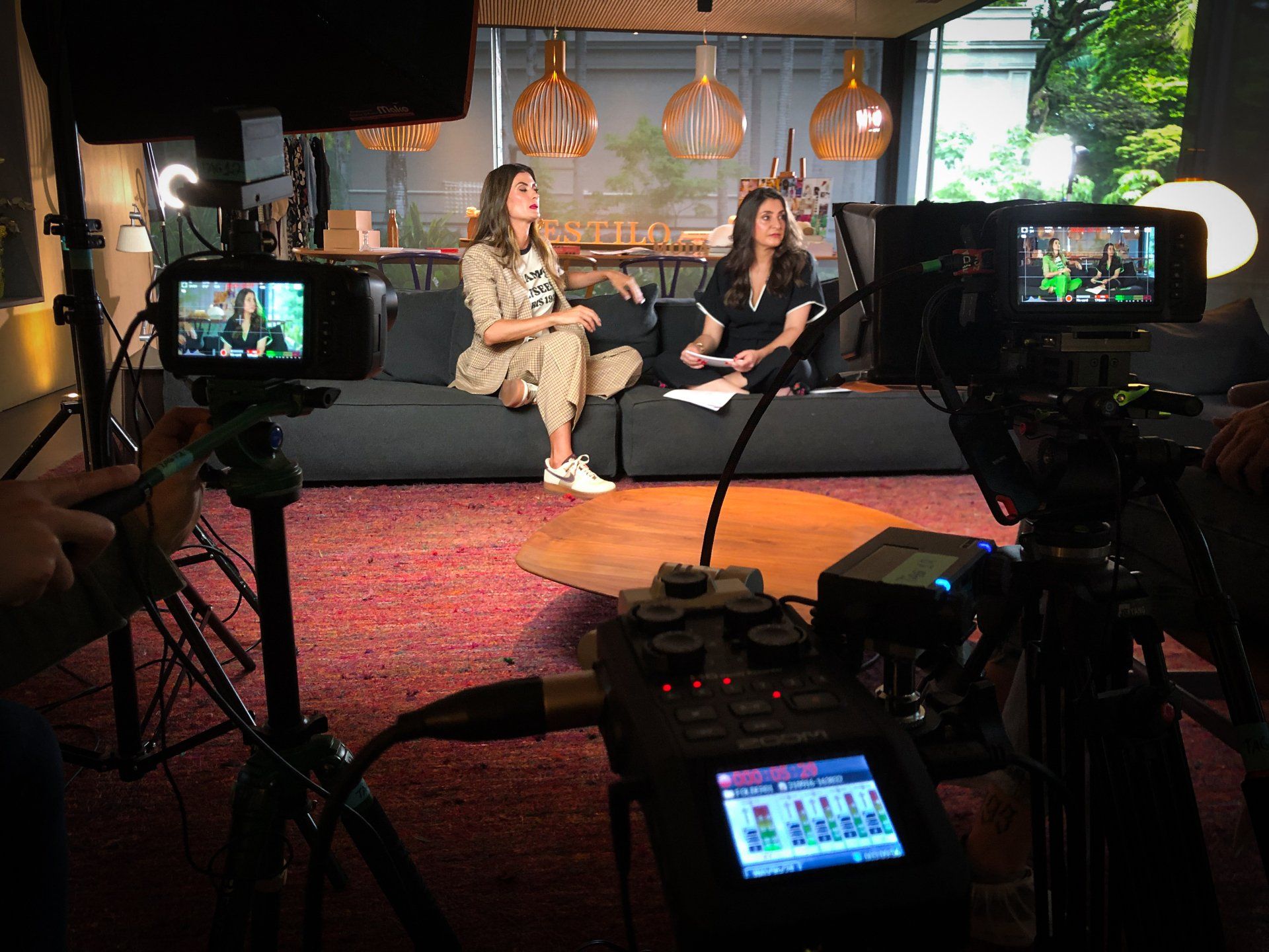 Duas mulheres sentadas em um sofá sendo filmadas em um estúdio com câmeras, luzes e equipamentos de áudio.