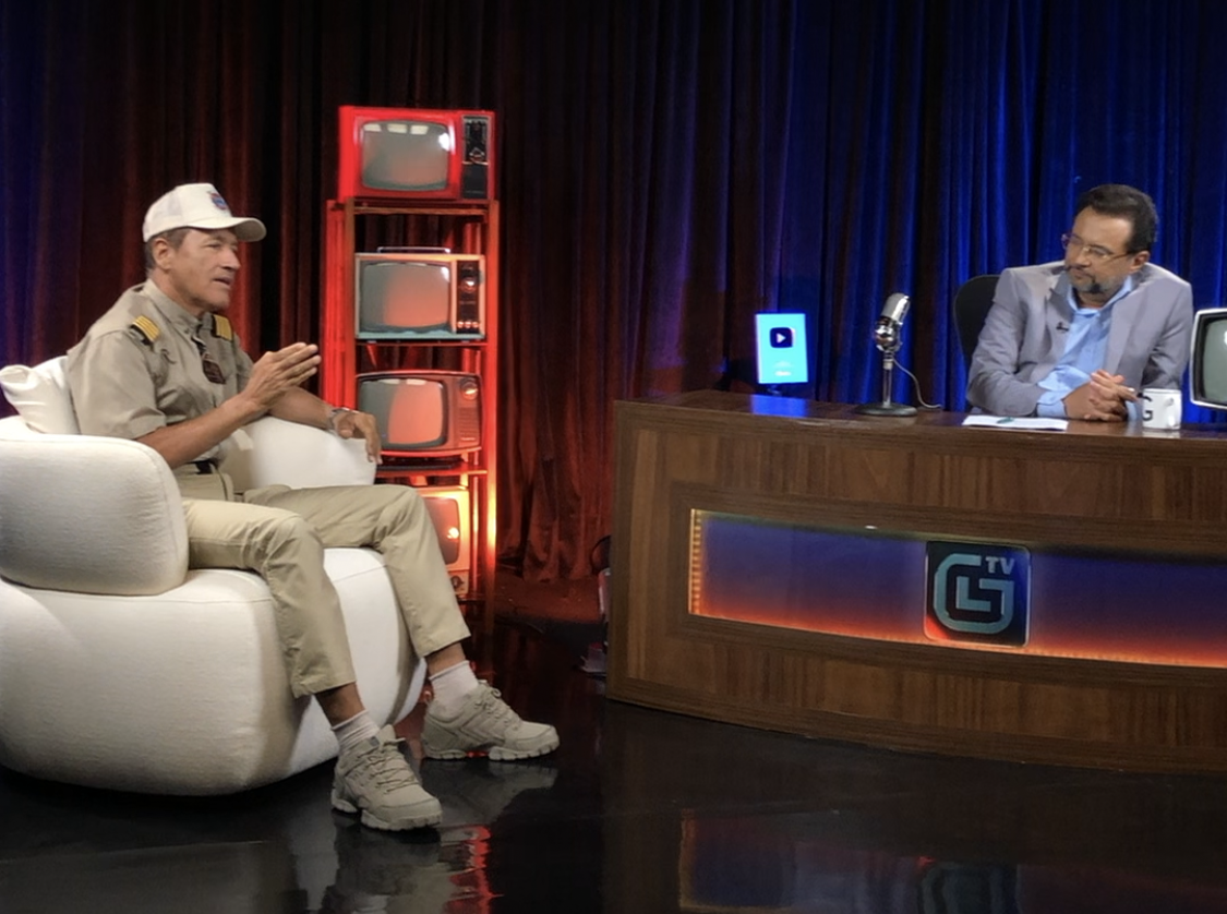 Talk show interview: guest in white cap speaks on couch while host sits at desk on stage.