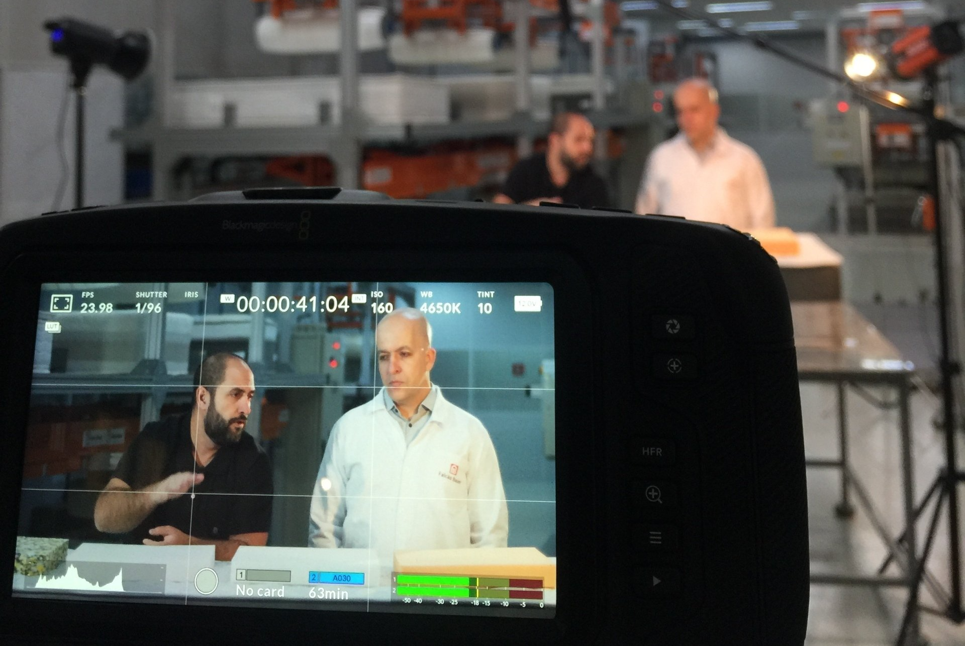 Uma câmera está gravando um vídeo de dois homens conversando em um laboratório de teste de colchão.