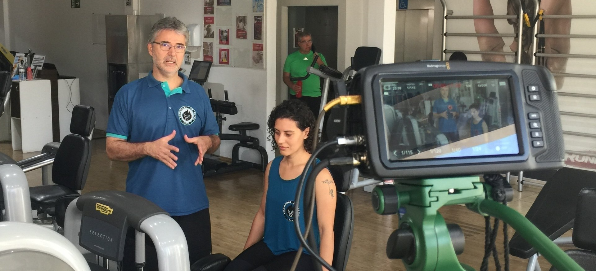 Um homem de camisa azul fala, uma mulher está sentada e uma câmera grava em uma academia. Outro homem está em pé atrás deles.