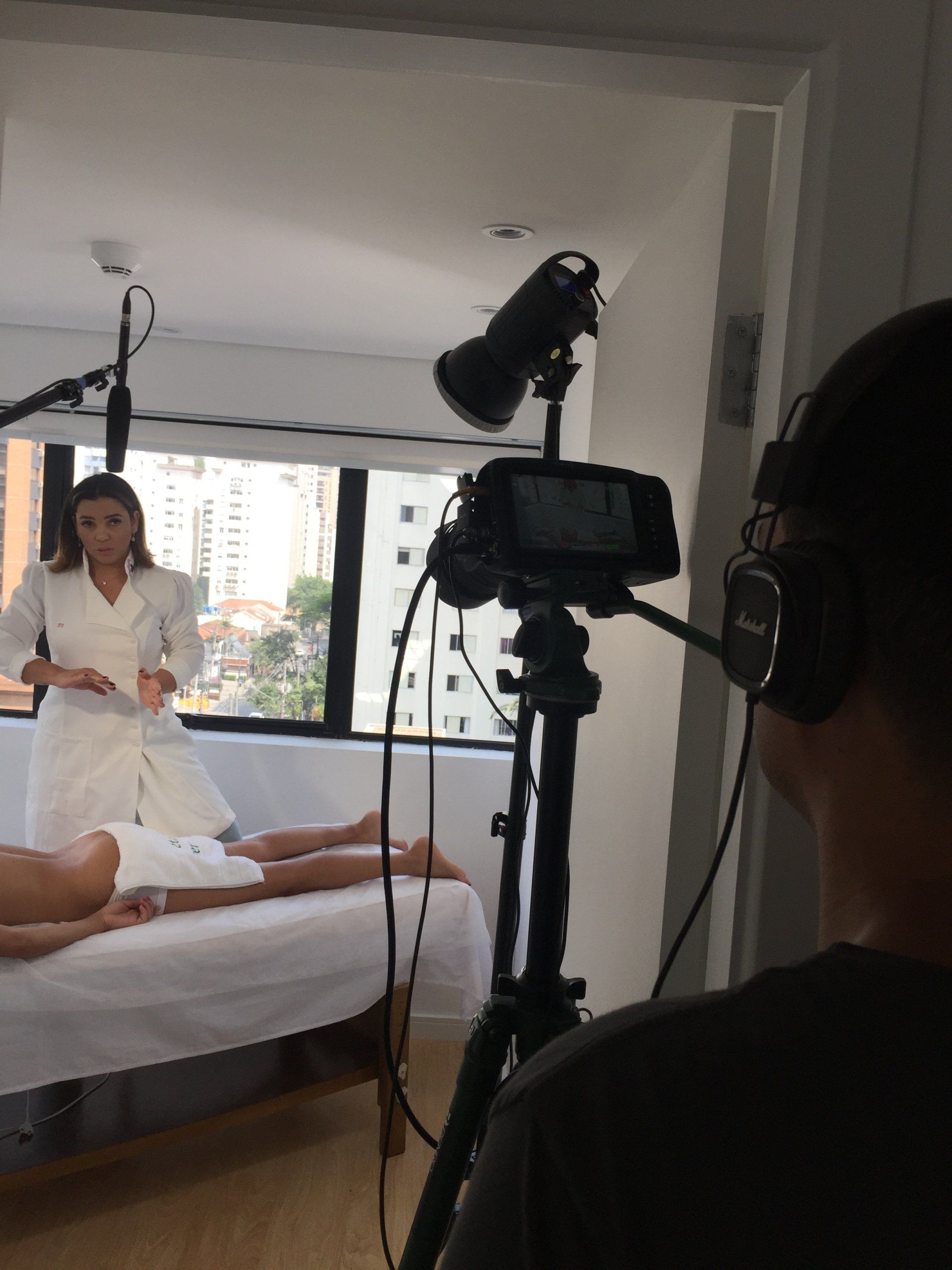 Uma pessoa está filmando uma mulher de jaleco branco perto de uma janela. 