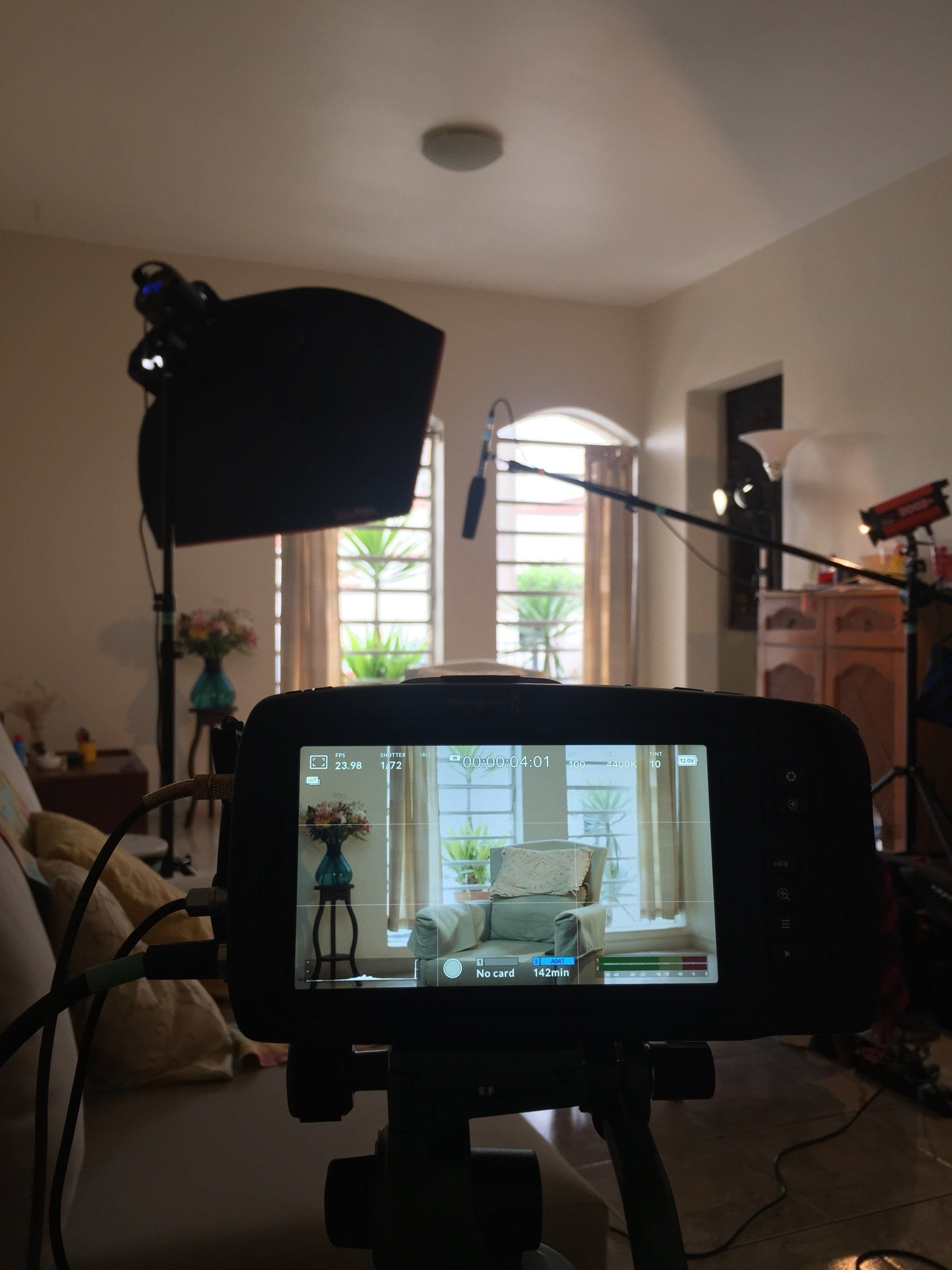 O visor de uma câmera mostra uma cena de sala de estar, com um microfone boom e luzes posicionadas para filmagem.