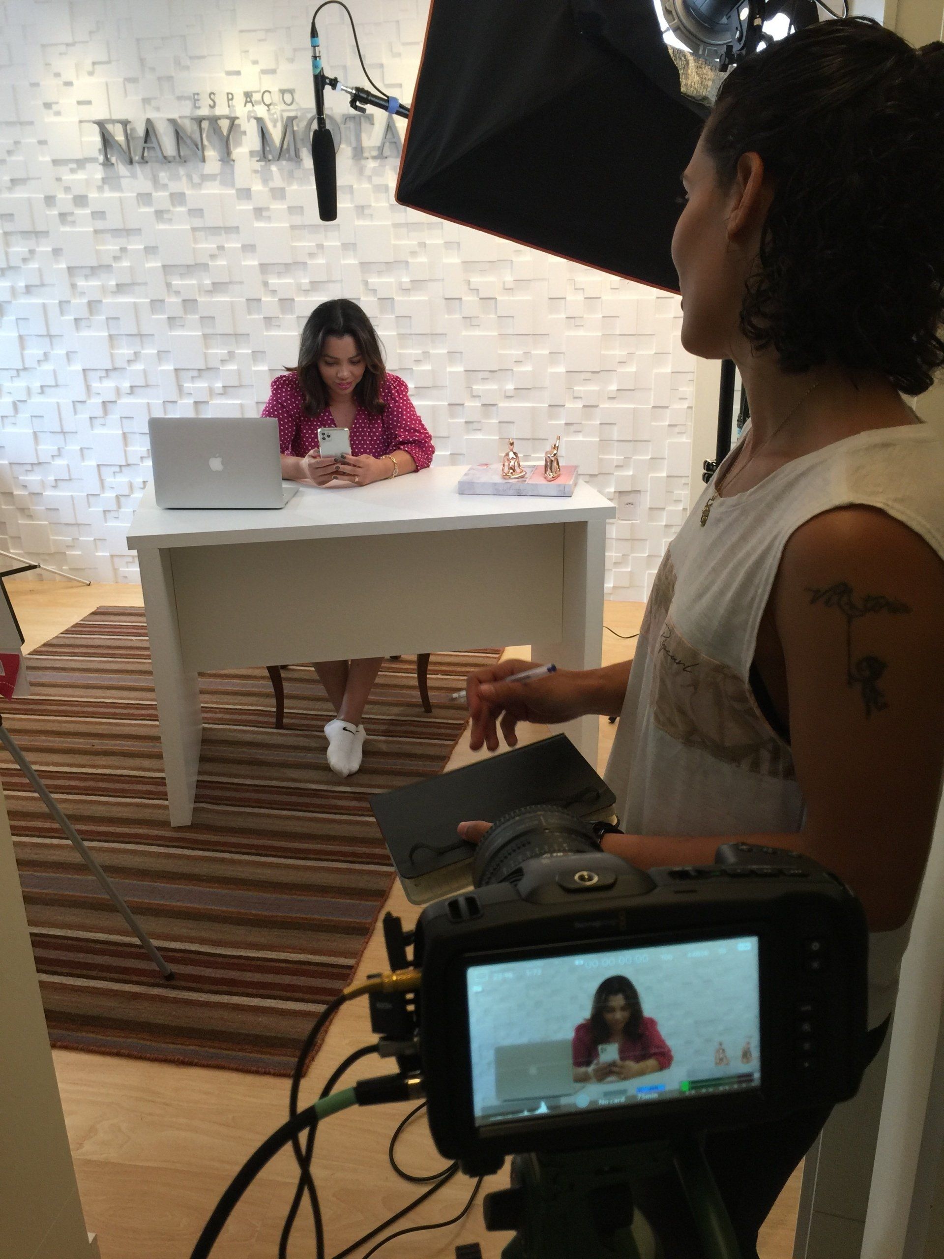 Mulher gravando vídeo, com laptop e luz softbox. Outra pessoa opera a câmera. Parede branca.
