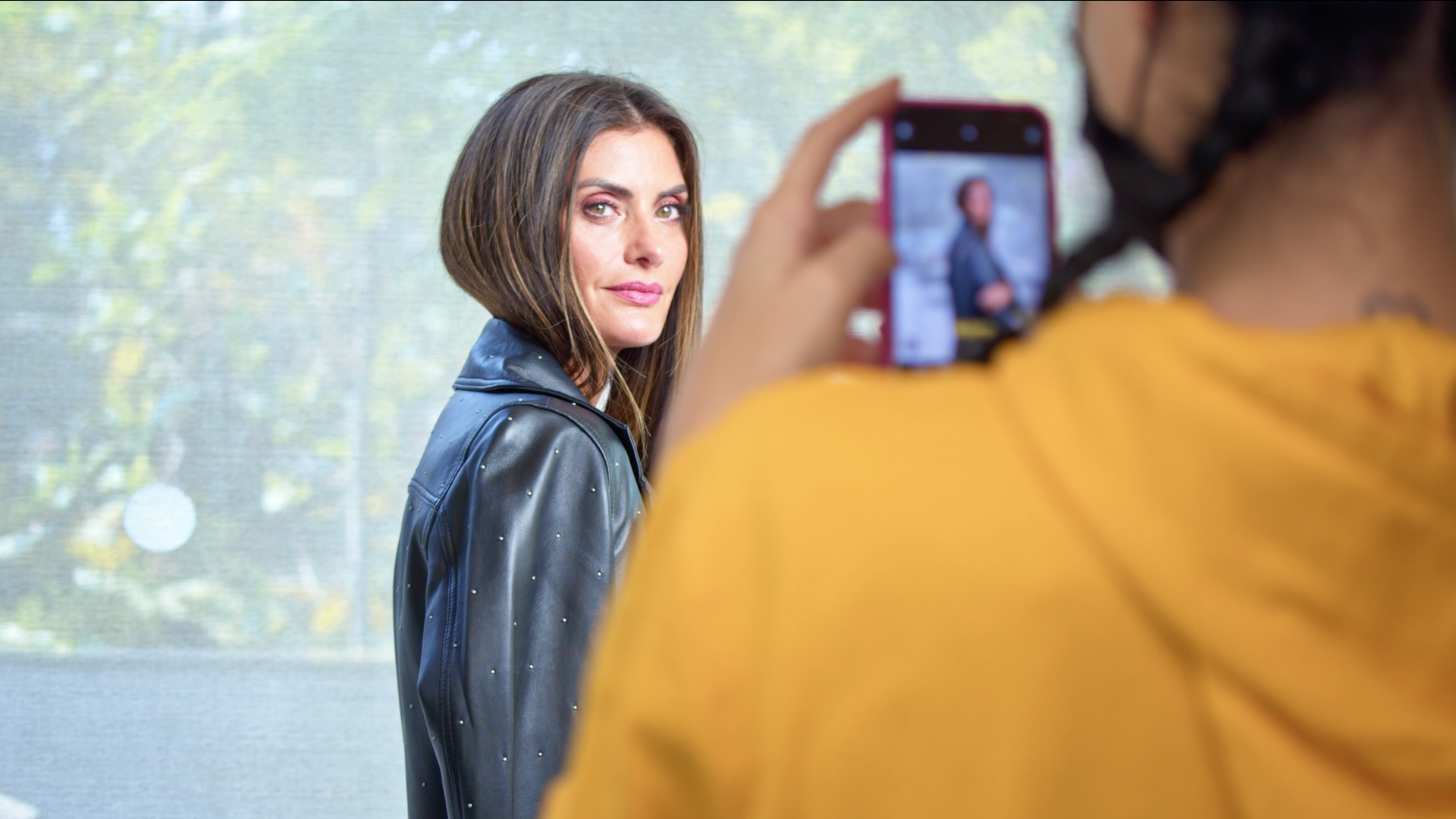 Mulher de jaqueta preta posa para foto tirada por pessoa usando máscara e camisa amarela.