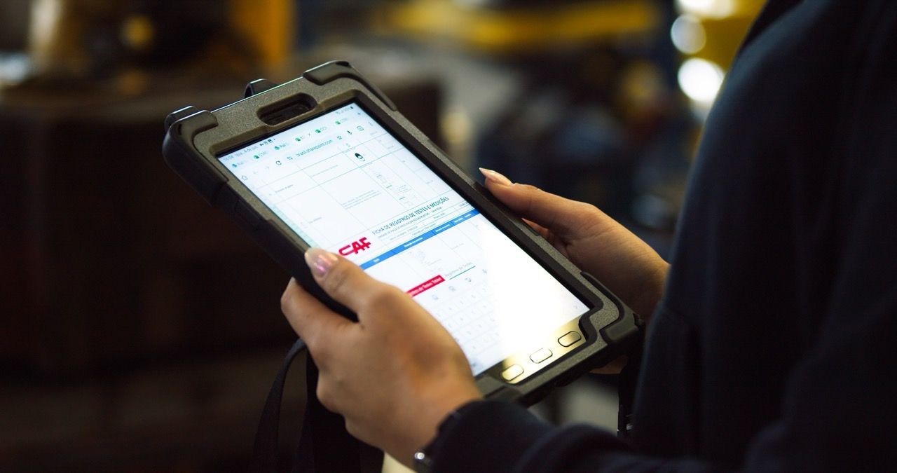 Hands holding a rugged tablet displaying text, likely for work, in a manufacturing setting.