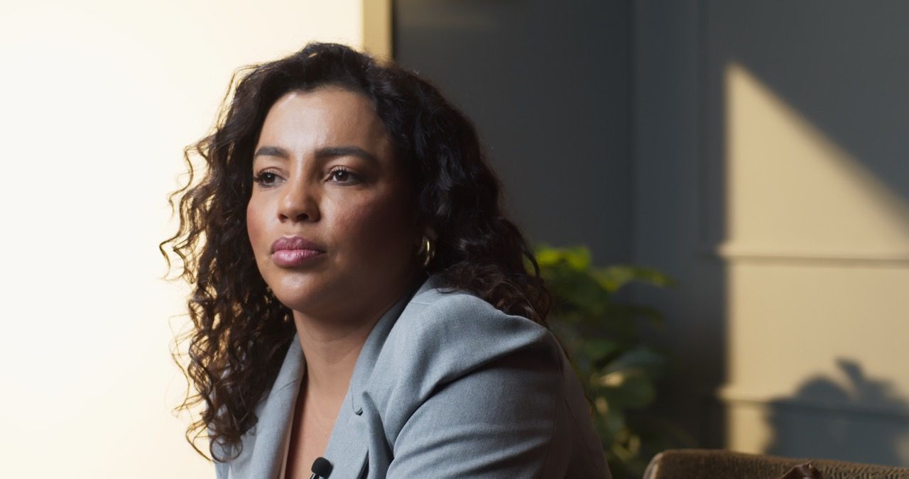 Uma mulher com cabelos cacheados está sentada em uma cadeira em frente a uma janela.