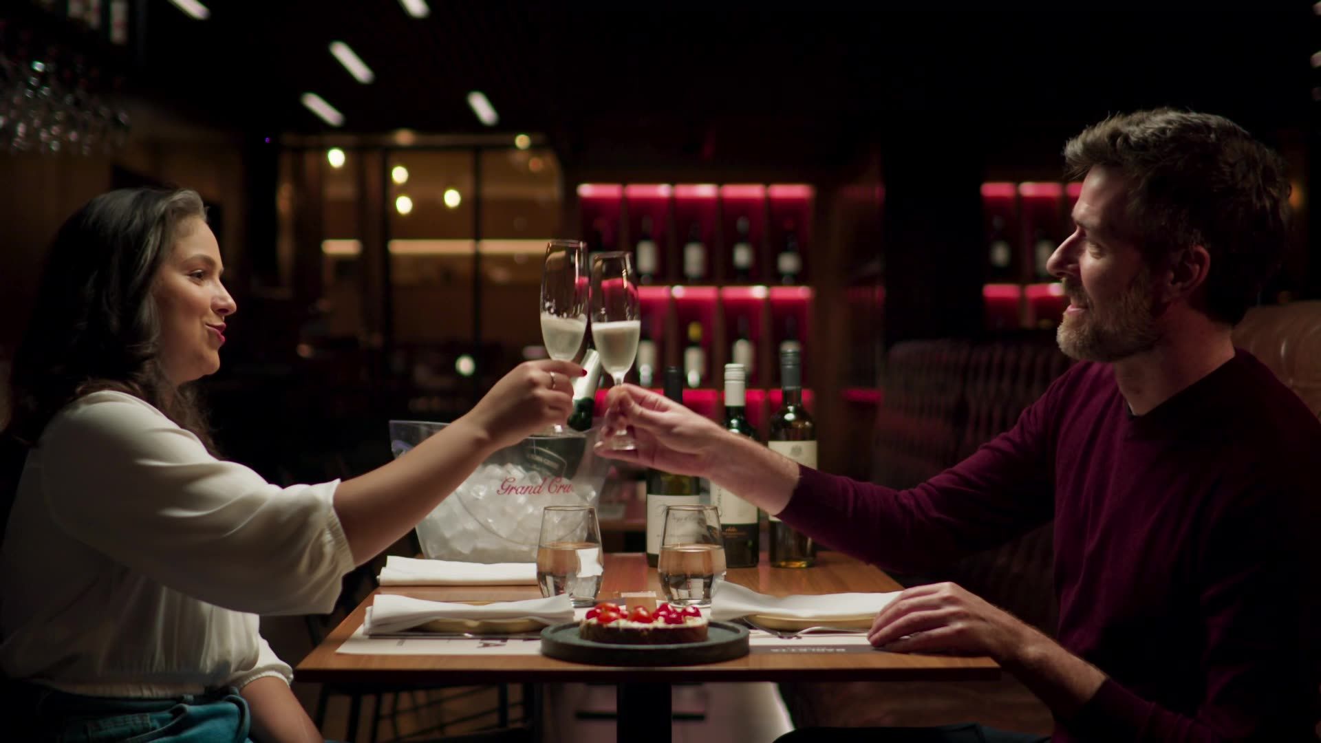 Casal brindando com taças de champanhe à mesa de um restaurante, sorrindo, em ambiente interno vermelho e escuro.