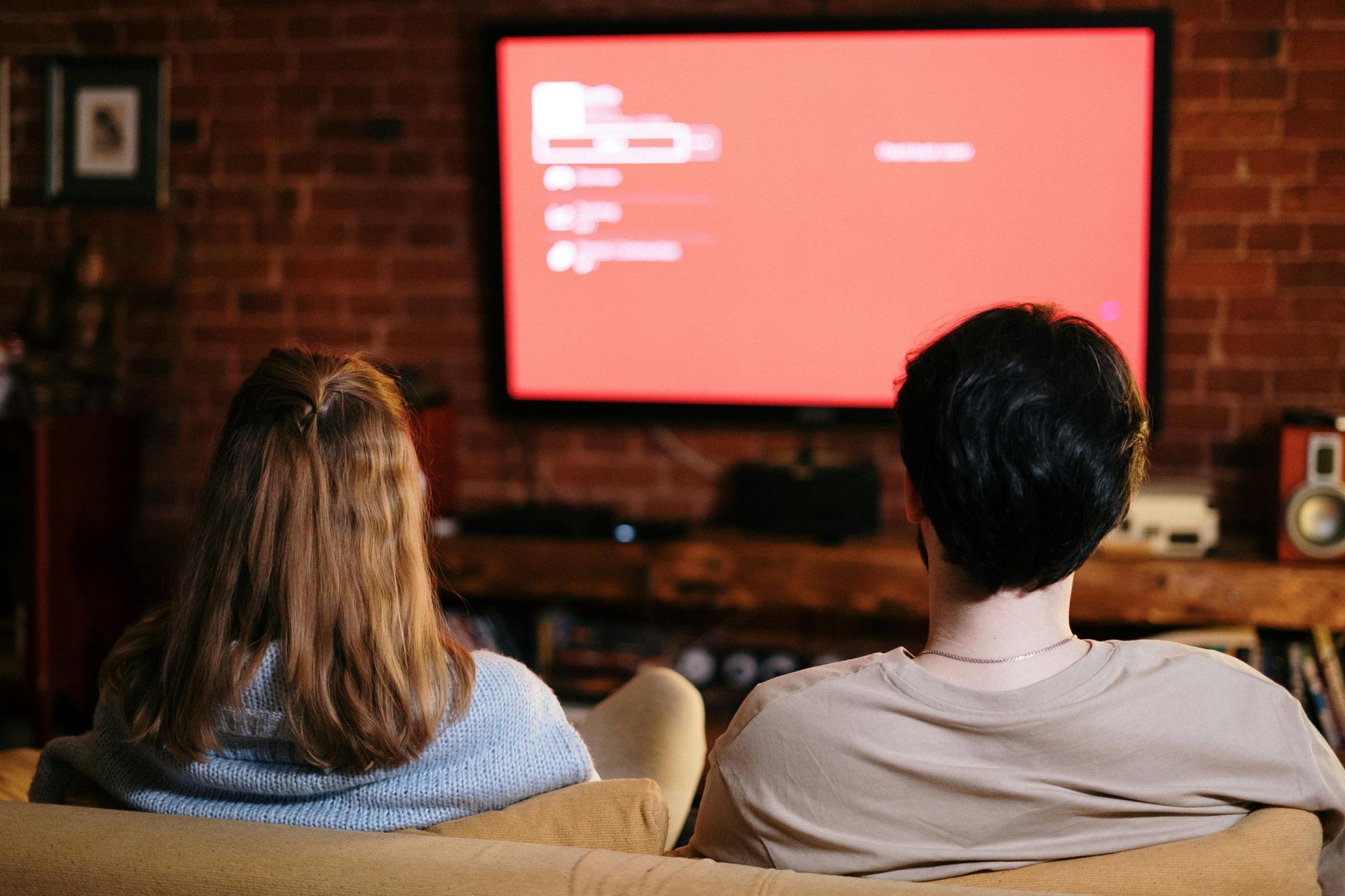 Two people sitting on a couch, facing a lit-up television in a brick-walled room.