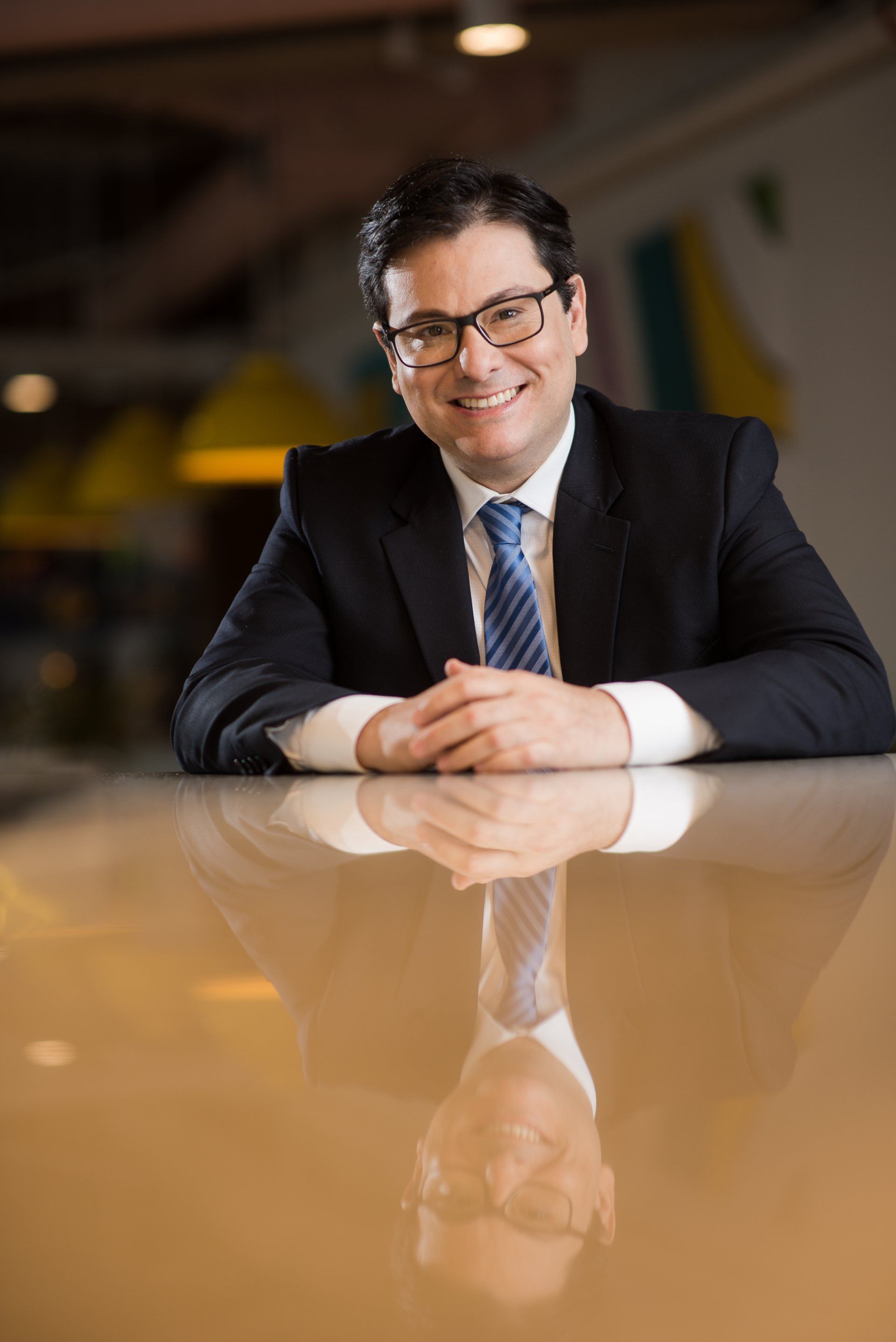 Um homem de terno sorri, apoiando os braços em uma superfície refletora.