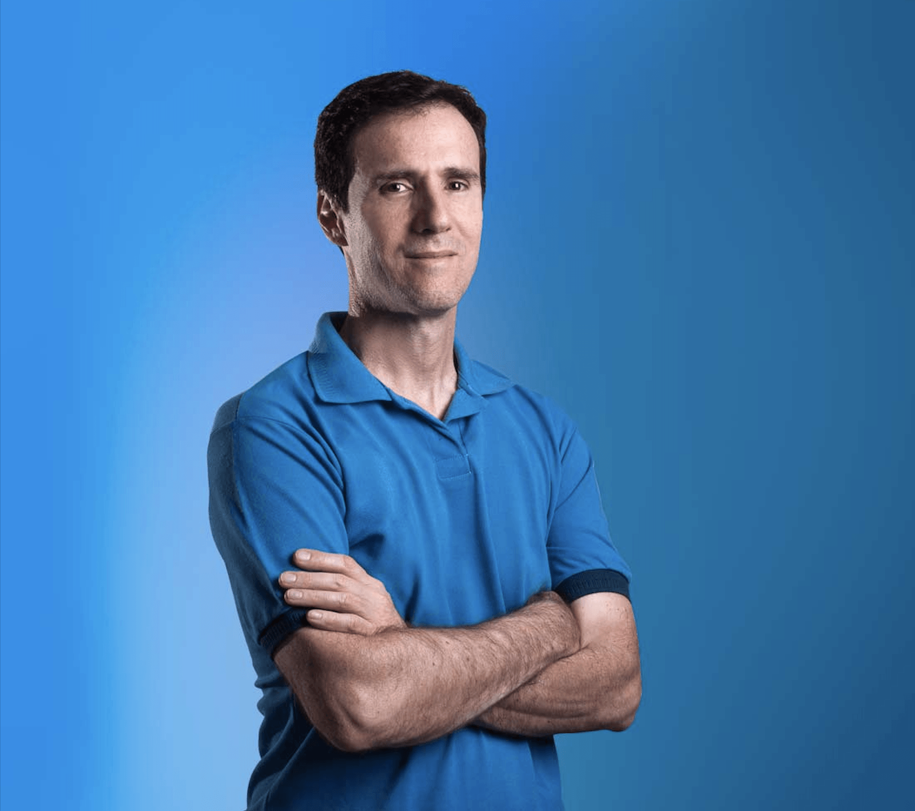 Homem com os braços cruzados, vestindo uma camisa polo azul contra um fundo azul degradê.