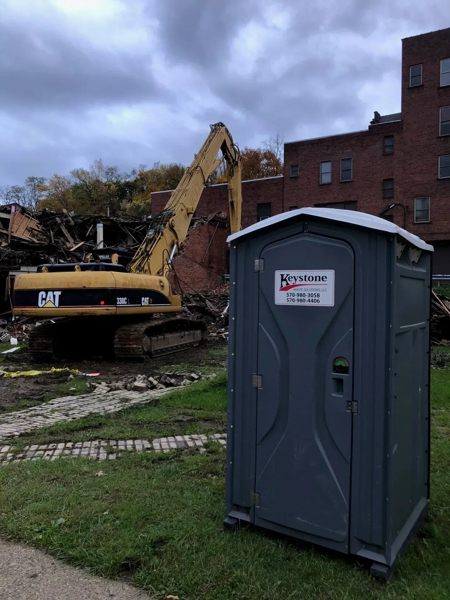 A portable toilet is sitting in front of a construction site with a cat excavator in the background.