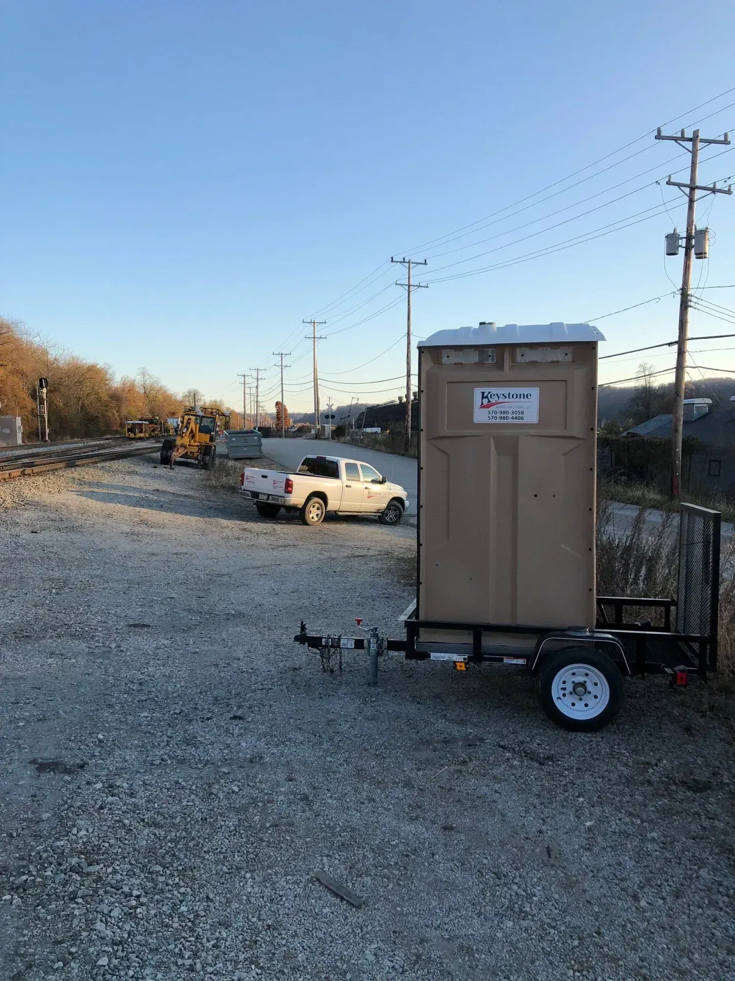 A trailer with a portable toilet attached to it is parked in a gravel lot.