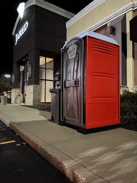 Red portable toilet next to a Jared jewelry store at night. A trash can is beside it on a concrete sidewalk.