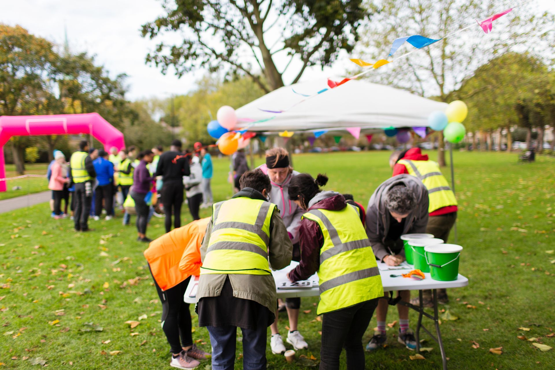 People registering at an outdoor event table in a park, wearing reflective vests. Pink archway, balloons, and flags are visible.