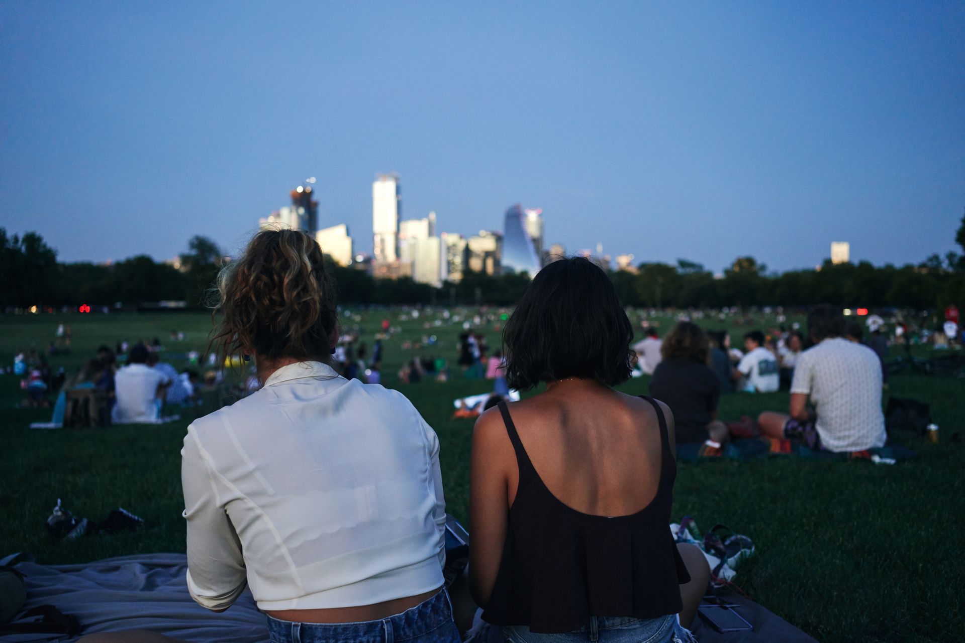 Two people sitting on a blanket in a park, looking toward a city skyline at dusk.