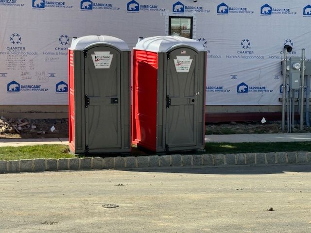 Two red and grey portable toilets sit outdoors in front of a building under construction.