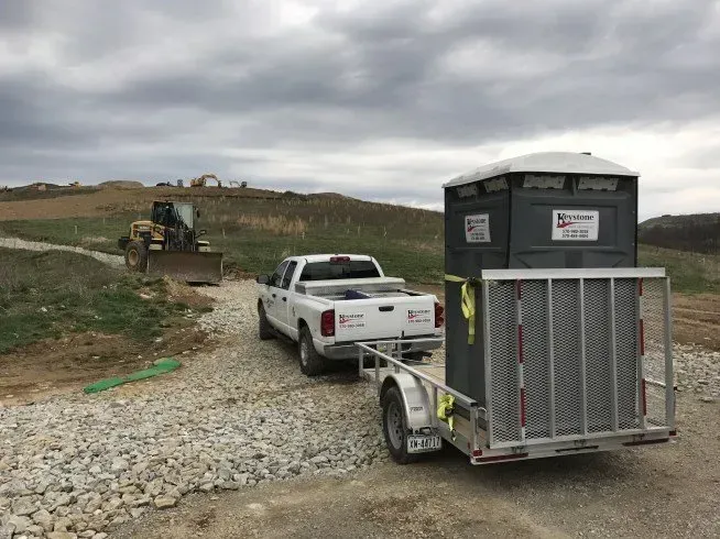 A white truck is towing a trailer with a portable toilet on it.