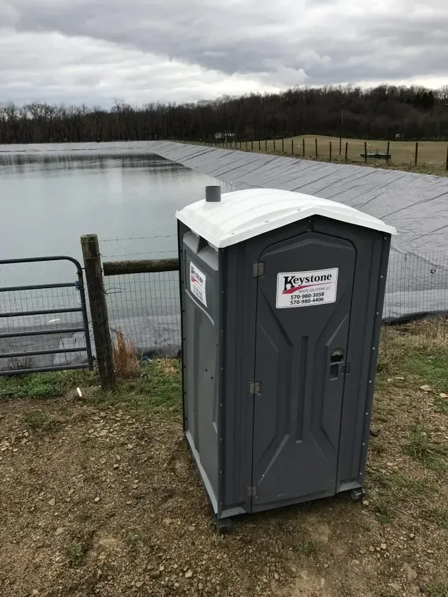 A portable toilet is sitting on top of a dirt field next to a lake.