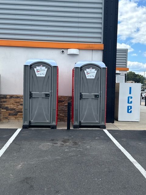 Two portable toilets on a paved lot beside a building. 'Ice' sign visible.