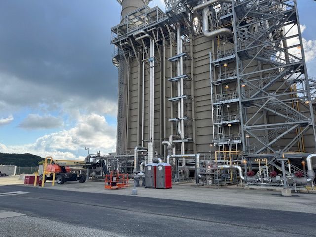 A large industrial power generation facility with tall steel structures and piping under a cloudy sky.