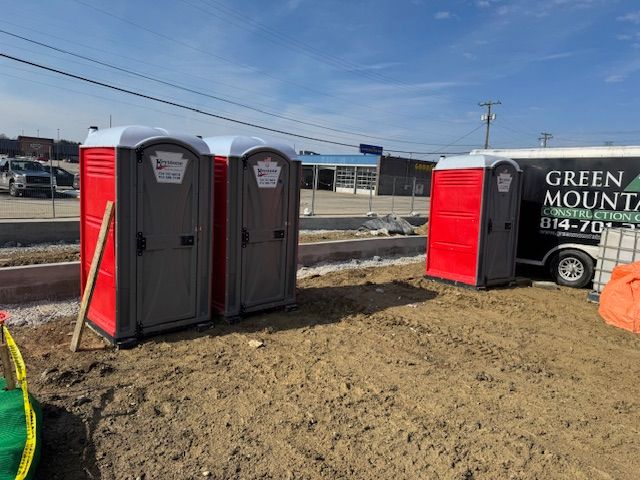 Red and gray portable toilets set up at an outdoor industrial work site. Three red and grey portable toilets sit on a dirt lot next to a Green Mountain construction trailer.