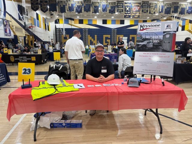 Vendor booth at indoor event with a red tablecloth, safety gear, and a seated man in a gymnasium.