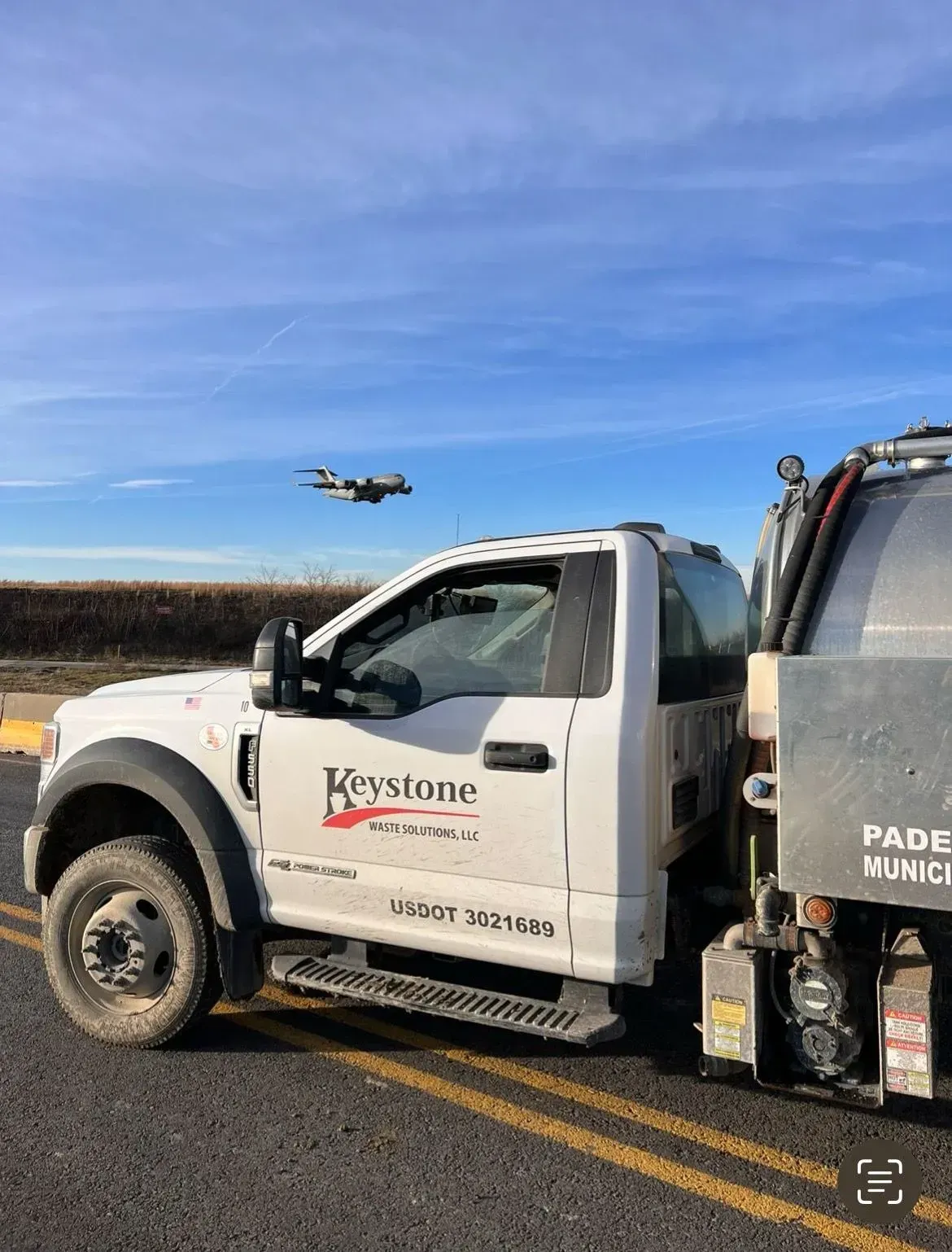 A white truck is parked on the side of the road with a plane flying in the background.