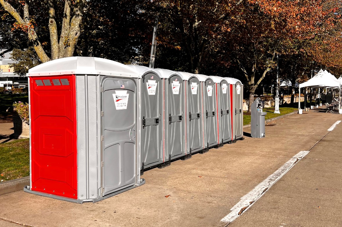 A row of portable toilets in a park; the first is red, the others gray.