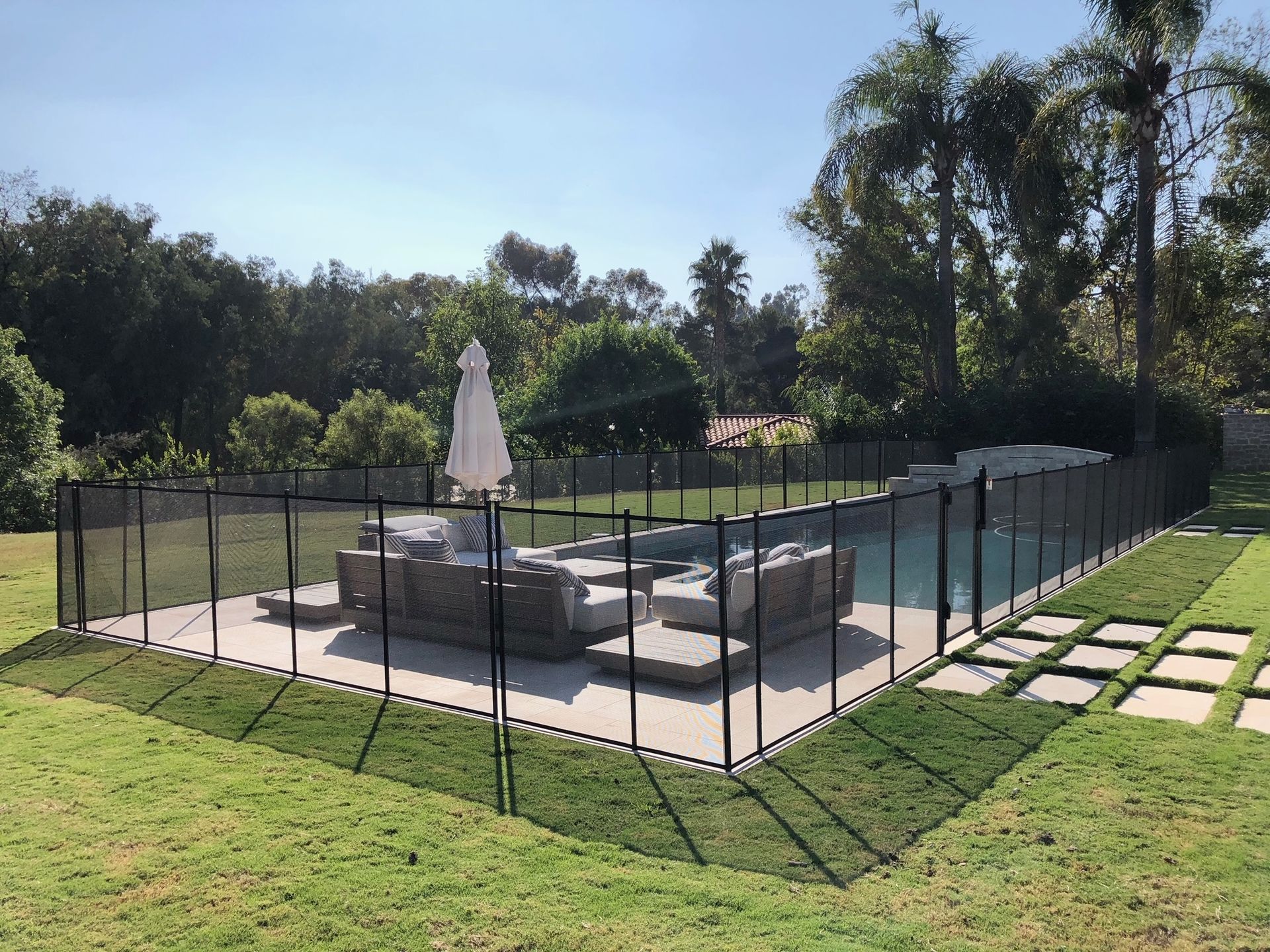 Pool enclosed by black mesh fence on green lawn, with patio furniture. Sunny day.