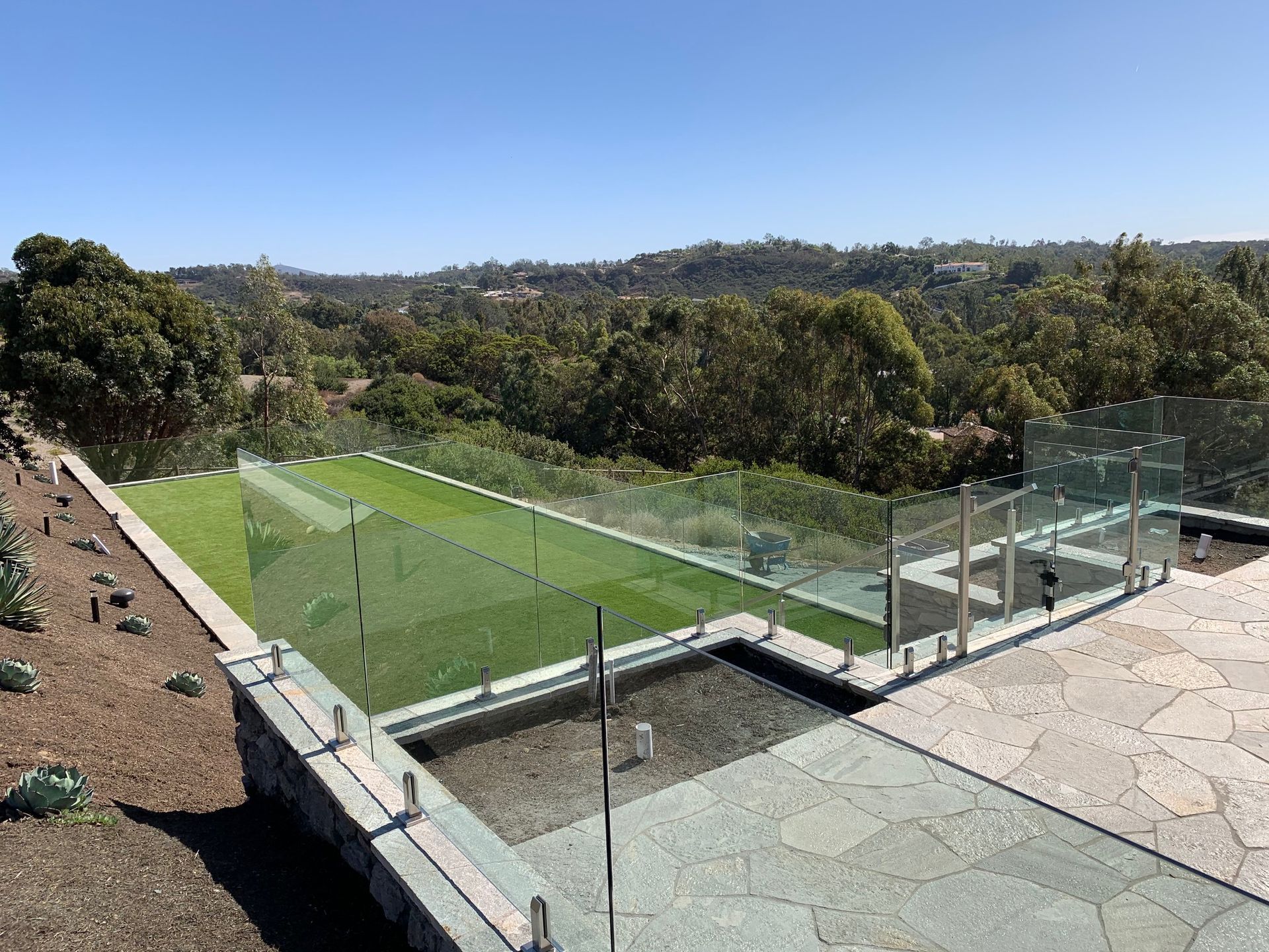 Glass-railed deck overlooking green lawn, water feature, and distant trees under blue sky.