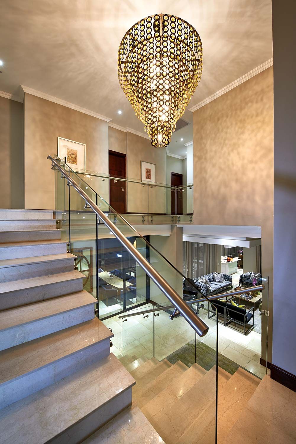 Staircase with glass railing, leading to a hallway with a large chandelier and a view of a living area below.
