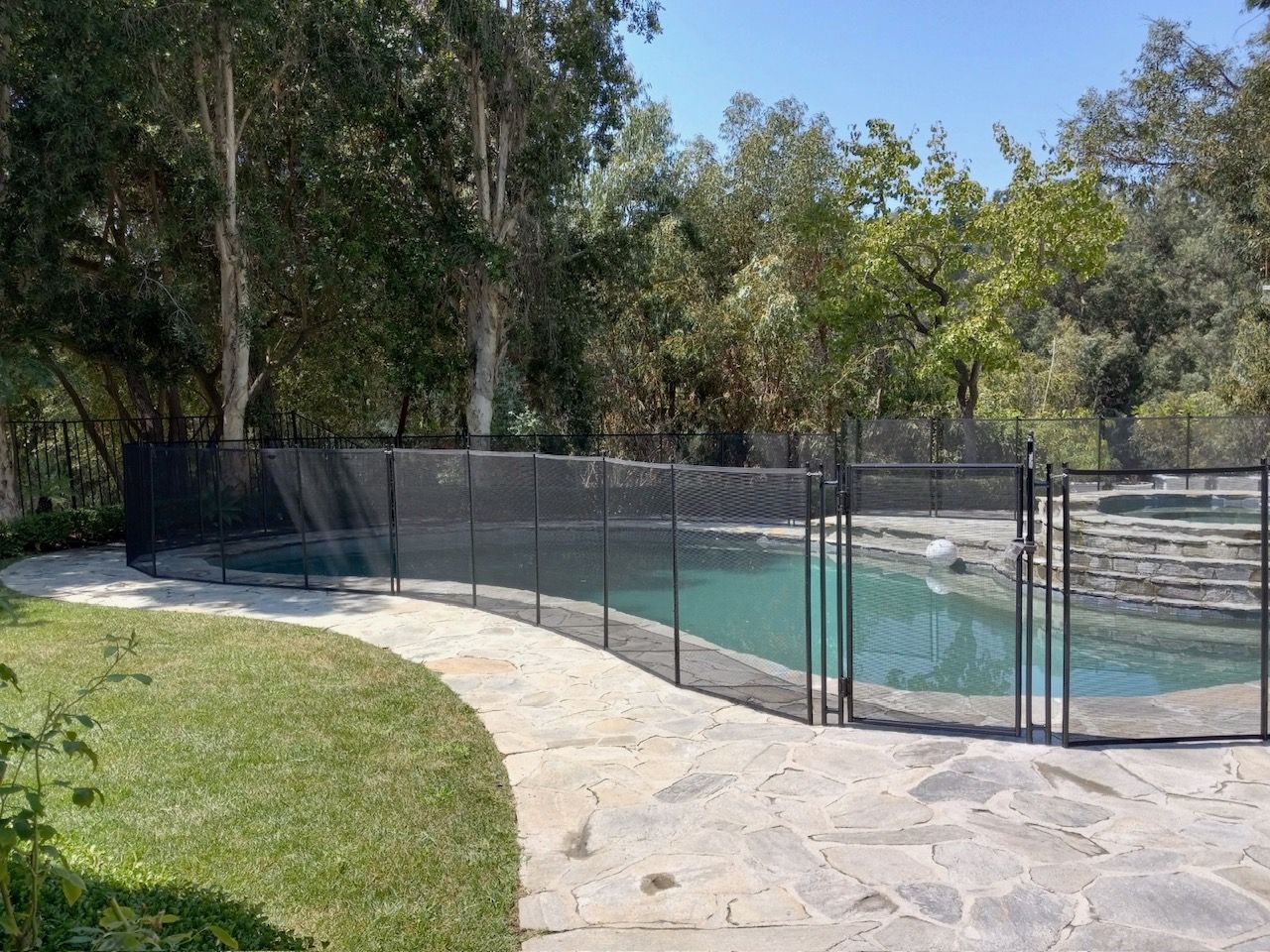 A pool surrounded by a black mesh safety fence on a stone patio, with green grass and trees in the background.